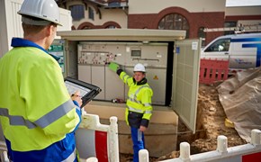 Zwei Techniker in Warnschutzjacken mit portablem Eingabegerät vor offener Trafostation Zwei Techniker in Warnschutzjacken mit portablem Eingabegerät vor offener Trafostation