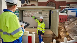 Zwei Techniker in Warnschutzjacken mit portablem Eingabegerät vor offener Trafostation Zwei Techniker in Warnschutzjacken mit portablem Eingabegerät vor offener Trafostation