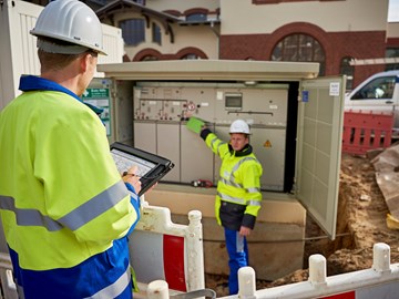 Zwei Techniker in Warnschutzjacken mit portablem Eingabegerät vor offener Trafostation Zwei Techniker in Warnschutzjacken mit portablem Eingabegerät vor offener Trafostation