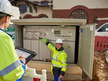 Zwei Techniker in Warnschutzjacken mit portablem Eingabegerät vor offener Trafostation Zwei Techniker in Warnschutzjacken mit portablem Eingabegerät vor offener Trafostation