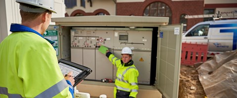 Zwei Techniker in Warnschutzjacken mit portablem Eingabegerät vor offener Trafostation Zwei Techniker in Warnschutzjacken mit portablem Eingabegerät vor offener Trafostation