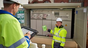 Zwei Techniker in Warnschutzjacken mit portablem Eingabegerät vor offener Trafostation Zwei Techniker in Warnschutzjacken mit portablem Eingabegerät vor offener Trafostation
