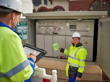 Zwei Techniker in Warnschutzjacken mit portablem Eingabegerät vor offener Trafostation Zwei Techniker in Warnschutzjacken mit portablem Eingabegerät vor offener Trafostation