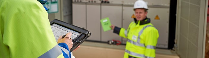 Zwei Techniker in Warnschutzjacken mit portablem Eingabegerät vor offener Trafostation Zwei Techniker in Warnschutzjacken mit portablem Eingabegerät vor offener Trafostation