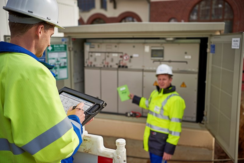Zwei Techniker in Warnschutzjacken mit portablem Eingabegerät vor offener Trafostation
