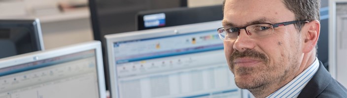Mitarbeiter am Schreibtisch vor zwei Monitoren schaut in die Kamera Mitarbeiter am Schreibtisch vor zwei Monitoren schaut in die Kamera