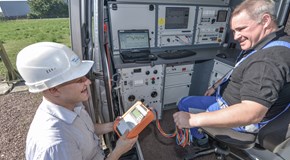 Lachender Arbeiter mit Helm und portablem Gerät vor einem offenen Technikfahrzeug, in dem ein Kollege sitzt Lachender Arbeiter mit Helm und portablem Gerät vor einem offenen Technikfahrzeug, in dem ein Kollege sitzt