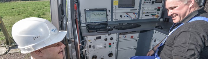 Lachender Arbeiter mit Helm und portablem Gerät vor einem offenen Technikfahrzeug, in dem ein Kollege sitzt Lachender Arbeiter mit Helm und portablem Gerät vor einem offenen Technikfahrzeug, in dem ein Kollege sitzt