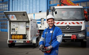 Mitarbeiter trägt Arbeitskleidung mit Helm und ein Bedienmodul um den Hals - er steht vor einem Lkw mit Kran und einem offenen Anhänger mit technischem Gerät Mitarbeiter trägt Arbeitskleidung mit Helm und ein Bedienmodul um den Hals - er steht vor einem Lkw mit Kran und einem offenen Anhänger mit technischem Gerät