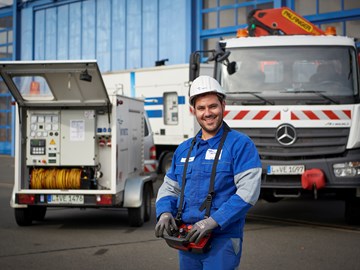 Mitarbeiter trägt Arbeitskleidung mit Helm und ein Bedienmodul um den Hals - er steht vor einem Lkw mit Kran und einem offenen Anhänger mit technischem Gerät Mitarbeiter trägt Arbeitskleidung mit Helm und ein Bedienmodul um den Hals - er steht vor einem Lkw mit Kran und einem offenen Anhänger mit technischem Gerät