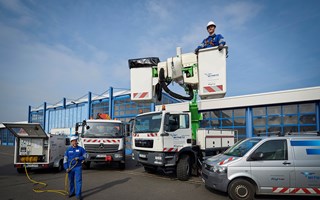 Mitarbeiter trägt Arbeitskleidung mit Helm und ein Bedienmodul um den Hals - er steht vor einem Lkw mit Kran und einem offenen Anhänger mit technischem Gerät Mitarbeiter trägt Arbeitskleidung mit Helm und ein Bedienmodul um den Hals - er steht vor einem Lkw mit Kran und einem offenen Anhänger mit technischem Gerät