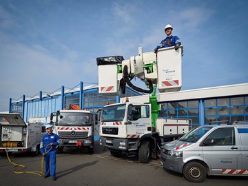 Auf einem Hof mit mehreren Fahrzeugen entrollt ein Mitarbeiter ein Kabel aus einem Anhänger, ein Kollege steht in der Kanzel eines Krans Auf einem Hof mit mehreren Fahrzeugen entrollt ein Mitarbeiter ein Kabel aus einem Anhänger, ein Kollege steht in der Kanzel eines Krans