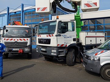 Auf einem Hof mit mehreren Fahrzeugen entrollt ein Mitarbeiter ein Kabel aus einem Anhänger, ein Kollege steht in der Kanzel eines Krans Auf einem Hof mit mehreren Fahrzeugen entrollt ein Mitarbeiter ein Kabel aus einem Anhänger, ein Kollege steht in der Kanzel eines Krans