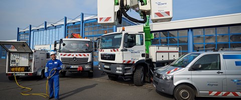 Auf einem Hof mit mehreren Fahrzeugen entrollt ein Mitarbeiter ein Kabel aus einem Anhänger, ein Kollege steht in der Kanzel eines Krans Auf einem Hof mit mehreren Fahrzeugen entrollt ein Mitarbeiter ein Kabel aus einem Anhänger, ein Kollege steht in der Kanzel eines Krans