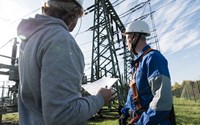 Zwei Männer mit weißen Helmen stehen unter Hochspannungsmasten, einer trägt eine Kletterausrüstung Zwei Männer mit weißen Helmen stehen unter Hochspannungsmasten, einer trägt eine Kletterausrüstung