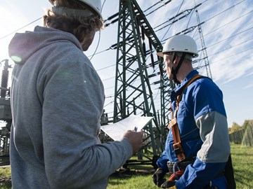 Zwei Männer mit weißen Helmen stehen unter Hochspannungsmasten, einer trägt eine Kletterausrüstung Zwei Männer mit weißen Helmen stehen unter Hochspannungsmasten, einer trägt eine Kletterausrüstung