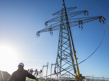 Der obere Teil eines Hochspannungsmasts hängt vor blauem Himmel an einem Kran, ein Arbeiter hält ein am Mast befestigtes Kabel Der obere Teil eines Hochspannungsmasts hängt vor blauem Himmel an einem Kran, ein Arbeiter hält ein am Mast befestigtes Kabel