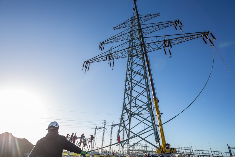 Der obere Teil eines Hochspannungsmasts hängt vor blauem Himmel an einem Kran, ein Arbeiter hält ein am Mast befestigtes Kabel