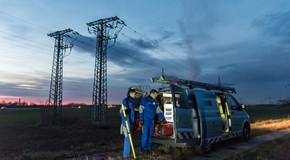 Zwei Techniker in blauen Overalls laden in der Dämmerung auf einem Feld mit Hochspannungsmasten Geräte aus einem Fahrzeug Zwei Techniker in blauen Overalls laden in der Dämmerung auf einem Feld mit Hochspannungsmasten Geräte aus einem Fahrzeug