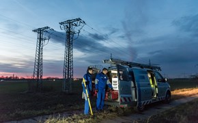 Zwei Techniker in blauen Overalls laden in der Dämmerung auf einem Feld mit Hochspannungsmasten Geräte aus einem Fahrzeug Zwei Techniker in blauen Overalls laden in der Dämmerung auf einem Feld mit Hochspannungsmasten Geräte aus einem Fahrzeug