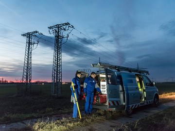 Zwei Techniker in blauen Overalls laden in der Dämmerung auf einem Feld mit Hochspannungsmasten Geräte aus einem Fahrzeug Zwei Techniker in blauen Overalls laden in der Dämmerung auf einem Feld mit Hochspannungsmasten Geräte aus einem Fahrzeug