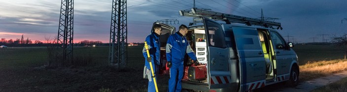 Zwei Techniker in blauen Overalls laden in der Dämmerung auf einem Feld mit Hochspannungsmasten Geräte aus einem Fahrzeug Zwei Techniker in blauen Overalls laden in der Dämmerung auf einem Feld mit Hochspannungsmasten Geräte aus einem Fahrzeug