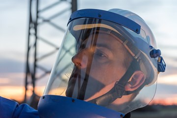 Monteur mit Helm und Schutzvisier vor einem Hochspannungsmast in der Dämmerung Monteur mit Helm und Schutzvisier vor einem Hochspannungsmast in der Dämmerung