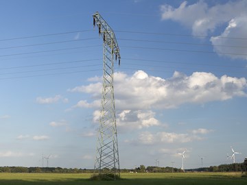 Hochspannungsmast auf einer grünen Wiese mit Windrädern unter blauem Himmel mit weißen Wolken Hochspannungsmast auf einer grünen Wiese mit Windrädern unter blauem Himmel mit weißen Wolken