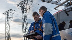 Zwei Monteure in Blaumännern blicken, am Heck eines Fahrzeugs vor zwei Hochspannungsmasten stehend, auf ein portables Eingabegerät Zwei Monteure in Blaumännern blicken, am Heck eines Fahrzeugs vor zwei Hochspannungsmasten stehend, auf ein portables Eingabegerät