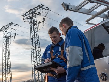 Zwei Monteure in Blaumännern blicken, am Heck eines Fahrzeugs vor zwei Hochspannungsmasten stehend, auf ein portables Eingabegerät Zwei Monteure in Blaumännern blicken, am Heck eines Fahrzeugs vor zwei Hochspannungsmasten stehend, auf ein portables Eingabegerät