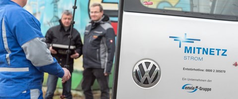 Zwei Männer in Parkern mit einem Arbeiter im Blaumann neben einer geöffneten Fahrzeugtür mit MITNETZ STROM-Logo Zwei Männer in Parkern mit einem Arbeiter im Blaumann neben einer geöffneten Fahrzeugtür mit MITNETZ STROM-Logo
