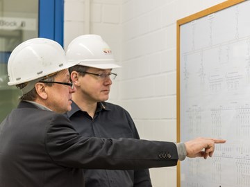 Zwei Männer mit weißen Helmen, der eine deutet mit ausgestrecktem Finger auf einen Plan an der Wand Zwei Männer mit weißen Helmen, der eine deutet mit ausgestrecktem Finger auf einen Plan an der Wand