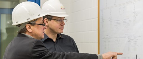 Zwei Männer mit weißen Helmen, der eine deutet mit ausgestrecktem Finger auf einen Plan an der Wand Zwei Männer mit weißen Helmen, der eine deutet mit ausgestrecktem Finger auf einen Plan an der Wand