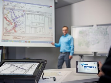 Laptop, portables Eingabegerät und Smartphone mit MITNETZ STROM-Logo - im Hintergrund hält ein Redner vor einem Plan seine Präsentation Laptop, portables Eingabegerät und Smartphone mit MITNETZ STROM-Logo - im Hintergrund hält ein Redner vor einem Plan seine Präsentation