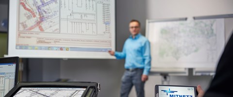 Laptop, portables Eingabegerät und Smartphone mit MITNETZ STROM-Logo - im Hintergrund hält ein Redner vor einem Plan seine Präsentation Laptop, portables Eingabegerät und Smartphone mit MITNETZ STROM-Logo - im Hintergrund hält ein Redner vor einem Plan seine Präsentation