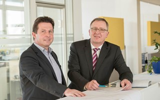 Dr. Schweer und Herr Hiersig an einem Counter über Unterlagen - blicken in die Kamera Dr. Schweer und Herr Hiersig an einem Counter über Unterlagen - blicken in die Kamera