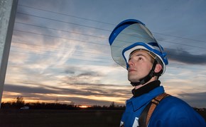 Monteur mit Helm und geöffnetem Schutzvisier vor einem Hochspannungsmast in der Dämmerung Monteur mit Helm und geöffnetem Schutzvisier vor einem Hochspannungsmast in der Dämmerung