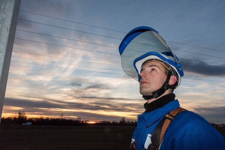 Monteur mit Helm und geöffnetem Schutzvisier vor einem Hochspannungsmast in der Dämmerung Monteur mit Helm und geöffnetem Schutzvisier vor einem Hochspannungsmast in der Dämmerung