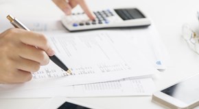 Hände mit Kugelschreiber und Taschenrechner über Unterlagen auf einem weißen Tisch Hände mit Kugelschreiber und Taschenrechner über Unterlagen auf einem weißen Tisch