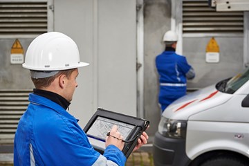 Zwei Techniker mit weißen Helmen und portablem Eingabegerät vor einer Anlage Zwei Techniker mit weißen Helmen und portablem Eingabegerät vor einer Anlage