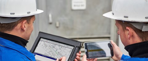 Zwei Techniker betrachten Pläne und Daten auf einem portablen Eingabegerät und einem Tablet nebeneinander Zwei Techniker betrachten Pläne und Daten auf einem portablen Eingabegerät und einem Tablet nebeneinander