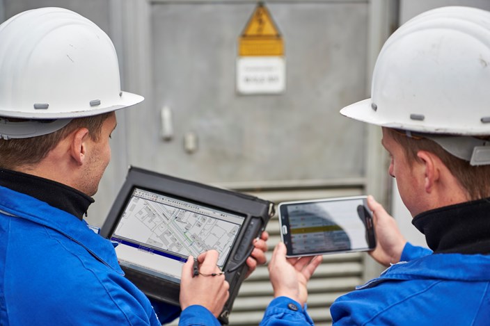 Zwei Techniker betrachten Pläne und Daten auf einem portablen Eingabegerät und einem Tablet nebeneinander Zwei Techniker betrachten Pläne und Daten auf einem portablen Eingabegerät und einem Tablet nebeneinander