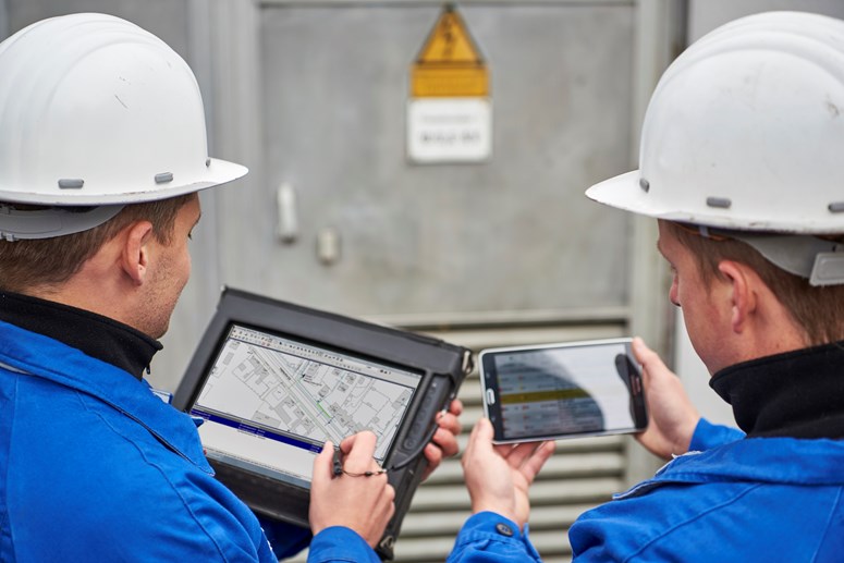 Zwei Techniker betrachten Pläne und Daten auf einem portablen Eingabegerät und einem Tablet nebeneinander Zwei Techniker betrachten Pläne und Daten auf einem portablen Eingabegerät und einem Tablet nebeneinander
