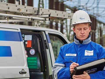 Mitarbeiter in blauer Arbeitskleidung mit Unterlagen vor einem Fahrzeug im Umspannwerk Mitarbeiter in blauer Arbeitskleidung mit Unterlagen vor einem Fahrzeug im Umspannwerk